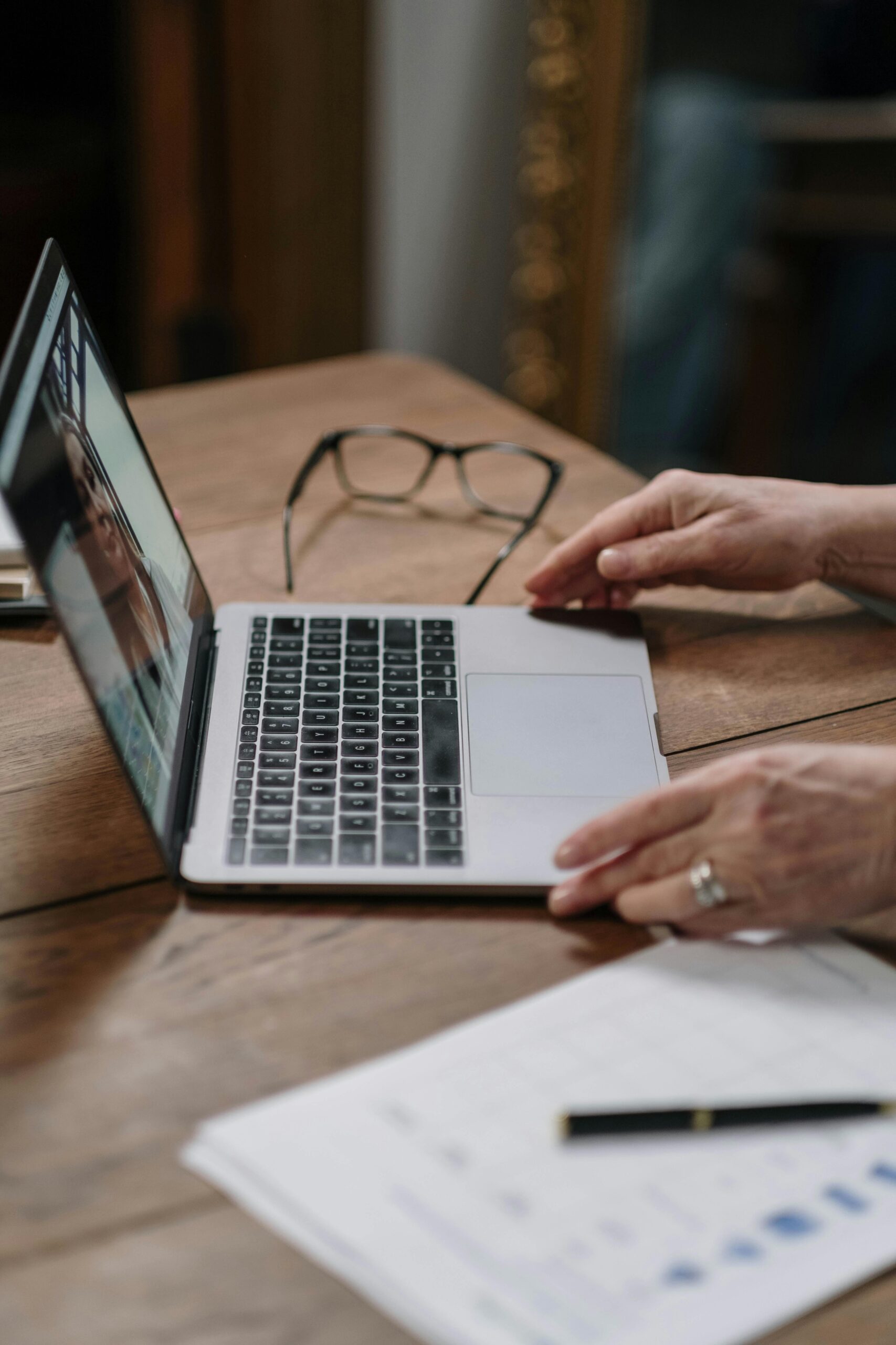 Hands on laptop keyboard with documents and eyeglasses on wooden desk.