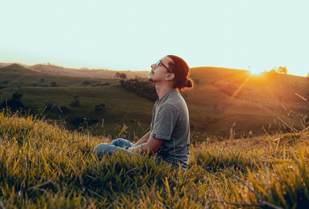 A man sits cross-legged in a wide, serene grassy field, deeply engaged in a meditative breathing exercise. His eyes are gently closed, his posture is upright but relaxed, and his hands rest comfortably on his knees. Soft, warm sunlight illuminates the natural landscape around him, highlighting the gentle sway of the grass and creating a peaceful, grounded atmosphere.